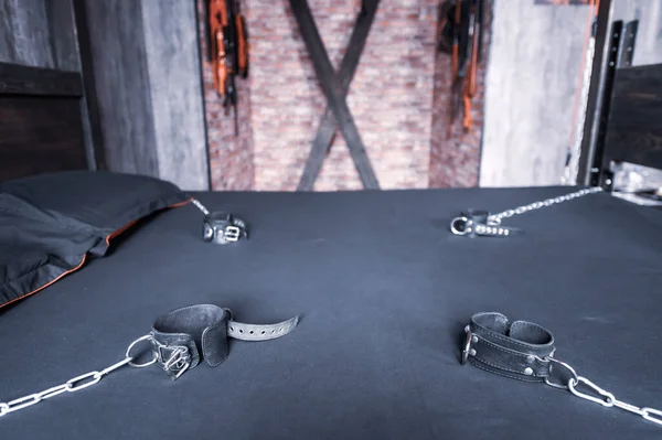 Torture of Andreev cross on the bedroom wall. VDSM equipment and sex toys. Leather handcuffs with a chain on a gray sheet. The lashes hang in the room for perversions. — Stock Photo, Image