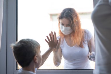 Sosyal mesafe. Tıbbi maskeli bir kadın oğluyla pencereden iletişim kuruyor. Dışardaki kız elini cam kapıya yasladı. Küçük çocuk izole edilmiş. Coronavirüs salgını.
