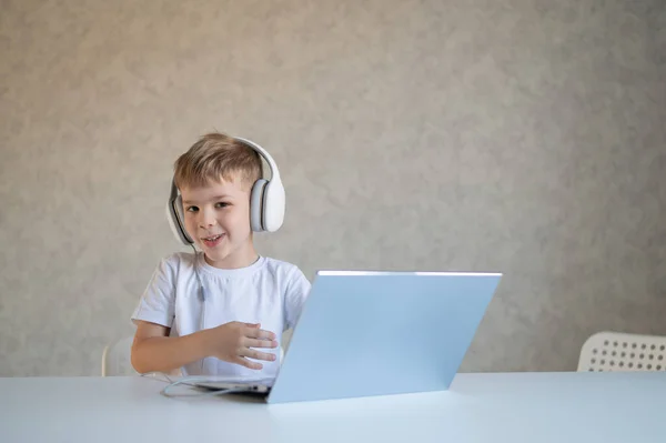Lindo chico europeo escucha música en los auriculares y los tipos en un ordenador portátil. El niño hace los deberes en la computadora. Un joven escolar recibe educación a distancia . — Foto de Stock