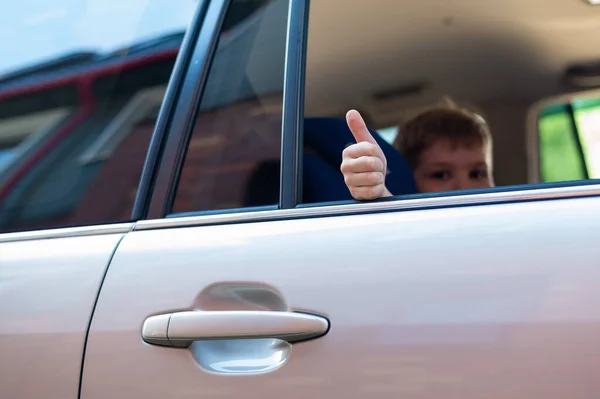 El niño se sienta en el asiento trasero de un coche plateado y muestra un pulgar. Preescolar está listo para un viaje emocionante . — Foto de Stock