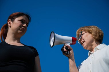 Nesillerin çatışması. Duygusal yaşlı bir kadın megafonla kızına bağırıyor. Yaşlı bir anne mavi arka planda hoparlörde orta yaşlı bir kadına küfrediyor..