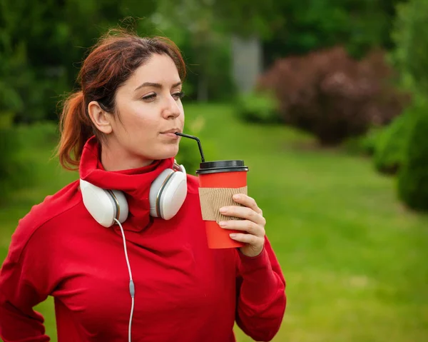 Red-haired woman with headphones and drinks coffee with a straw in the park. Girl enjoys a walk on a sunny summer day and holds a takeaway cappuccino cup. Headphones connected to a smartphone.