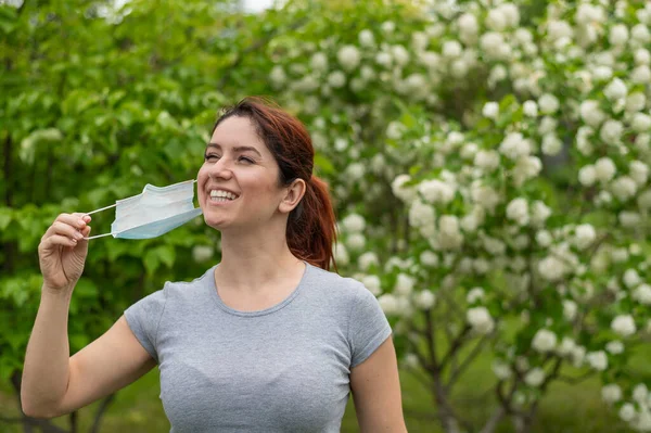 Uma mulher com uma t-shirt cinzenta tira alegremente a máscara médica no final da quarentena. Uma menina celebra o fim da quarentena enquanto caminha em um parque. Respiração livre sem alergias. — Fotografia de Stock