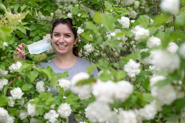 Eine fröhliche rothaarige Frau zieht eine medizinische Maske ab, während sie neben einem blühenden Apfelbaum in einem Park steht. Beendigung der saisonalen allergischen Rhinitis. Das Mädchen wurde von einer Pollenallergie geheilt. — Stockfoto