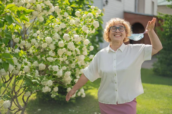 Щаслива стара жінка знімає маску і глушить з квітучої яблуні. Жінка-пенсіонер насолоджується свіжим повітрям у теплий літній день у саду. Карантин закінчився. Кінець пандемії коронавірусу . — стокове фото