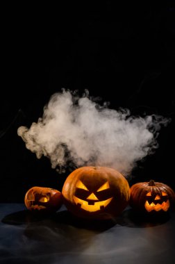 Siyah arka planda oyulmuş parlak bir sırıtışla üç ürkütücü cadılar bayramı buharı kabağı. El yapımı bir kriko-fener kafası ve içinde sis arasında karanlıkta bir mum. Şeker mi şaka mı? Kartpostal.