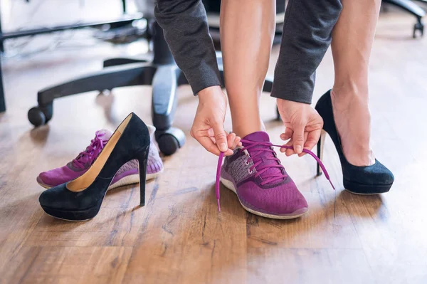 Κοντινό πλάνο ποδιών μιας υπαλλήλου γραφείου με πόνο στα πόδια. Μια γυναίκα αλλάζει ψηλά μαύρα τακούνια σουέτ σε πιο άνετα παπούτσια. Το κορίτσι βγάζει τα παπούτσια της και φοράει μωβ αθλητικά παπούτσια.. — Φωτογραφία Αρχείου