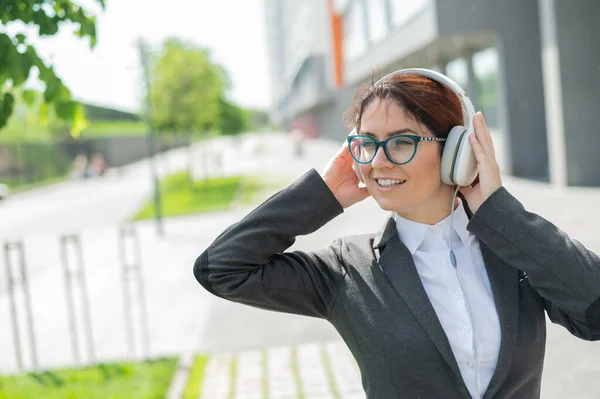 Porträtt av en leende kvinna i kostym som lyssnar på musik i hörlurar utomhus. Kvinnlig kontorsanställd håller händerna på hörlurar njuter sång och dans på gatan. — Stockfoto
