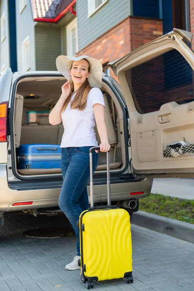Eine Europäerin mit Hut packt allein ihre Koffer für eine Autofahrt. Schöne glückliche Mädchen lädt einen Koffer in den Kofferraum. Gelbes Gepäck für die Sommerferien. — Stockfoto