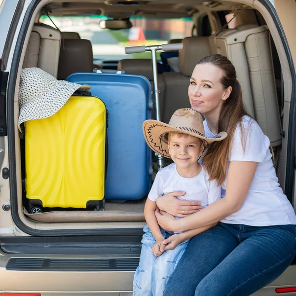 Europäerin sitzt mit ihrem Sohn im Kofferraum eines Geländewagens. Schöne glückliche Mutter liebevoll umarmt Sohn. Gelbes Gepäck für den Sommerurlaub mit dem Auto. Urlaub mit der ganzen Familie. — Stockfoto