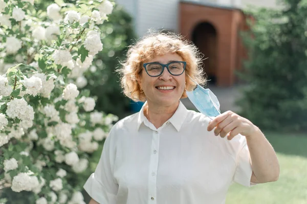 Glad gammal kvinna tar av masken och sniffar på ett blommande äppelträd. En kvinnlig pensionär njuter av frisk luft en varm sommardag i trädgården. Karantänen är över. Slutet på coronaviruspandemin. — Stockfoto