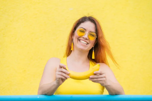 Красивая рыжая женщина в солнечных очках держит в руках банан и дураков. Привлекательная европейская девушка в платье позирует на желтом фоне очаровательно улыбается. — стоковое фото