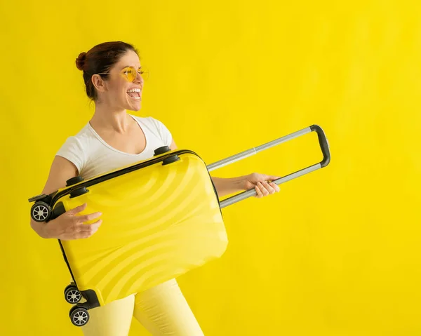 Smiling woman in sunglasses fooling around and holding a suitcase like a guitar on a yellow background. An excited girl in anticipation of a summer vacation trip simulates playing a string instrument. — Stock Photo, Image