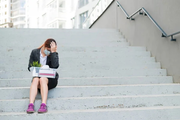 Una mujer con una caja de pertenencias personales se sienta en las escaleras y dobla las manos desesperada en la cabeza. Empleada con máscara médica despedida debido a crisis financieras en coronovirus. Desempleo —  Fotos de Stock