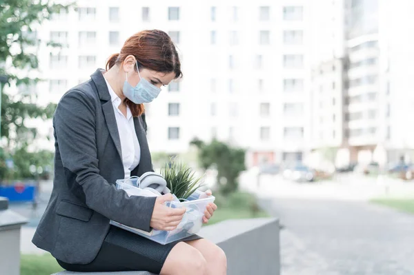 Mujer deprimida en máscara médica se sienta en el parapeto al aire libre con una caja de artículos personales desde el escritorio. Una chica desempleada sola en la calle. Quiebra de una empresa por cuarentena en coronavirus. —  Fotos de Stock