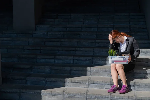 Mujer deprimida vestida con máscara médica es despedida y está sentada en las escaleras con una caja de pertenencias personales. Trabajadora de oficina con traje y zapatillas al aire libre. El desempleo en la crisis económica —  Fotos de Stock