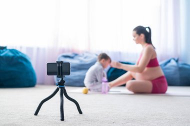 Hamile bir kadın oğluyla jimnastik yapıyor. Müstakbel anne telefonda blogu için bir eğitim videosu çekiyor. Bebekli fitness.