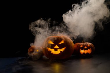 Siyah arka planda oyulmuş parlak bir sırıtışla üç ürkütücü cadılar bayramı buharı kabağı. El yapımı bir kriko-fener kafası ve içinde sis arasında karanlıkta bir mum. Şeker mi şaka mı? Kartpostal.