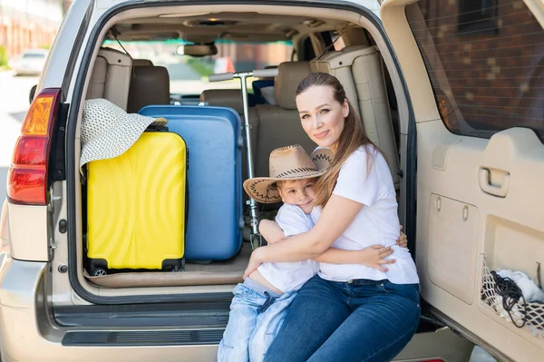 Europäerin sitzt mit ihrem Sohn im Kofferraum eines Geländewagens. Schöne glückliche Mutter liebevoll umarmt Sohn. Gelbes Gepäck für den Sommerurlaub mit dem Auto. Urlaub mit der ganzen Familie. — Stockfoto