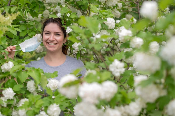 Eine fröhliche rothaarige Frau zieht eine medizinische Maske ab, während sie neben einem blühenden Apfelbaum in einem Park steht. Beendigung der saisonalen allergischen Rhinitis. Das Mädchen wurde von einer Pollenallergie geheilt. — Stockfoto