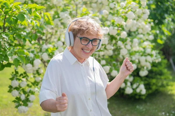 Glad gammal kvinna lyssnar på musik på hörlurar och danser i en park av blommande äppelträd. Aktiva kvinnliga pensionärer ler och njuter av en varm sommardag i trädgården. Avancerad äldre generation. — Stockfoto