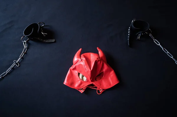 Equipamento BDSM. Vista superior de uma máscara de couro vermelho diabo e algemas pretas com correntes em uma folha cinza. Brinquedos sexuais. Dominância. Não há pessoas . — Fotografia de Stock