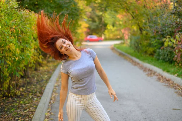 Uma bela mulher ruiva está andando pela rua e dançando. Menina feliz em uma camiseta cinza e leggings listrados caminha e se diverte no parque de outono. Foto em movimento. — Fotografia de Stock