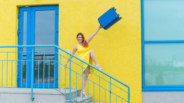 Šťastná zrzavá žena ve žlutých šatech sestupuje ze schodů a radostně houpe modrým kufříkem. Dívka se připravuje na cestu. Koncept letních prázdnin. — Stock fotografie