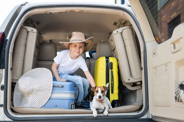 Glücklicher Junge mit Cowboyhut und Welpe Jack Russell Terrier reisen mit dem Auto. Ein Kind und ein lustiger kleiner Hund sitzen im Kofferraum und sind bereit für die Sommerferien. Unabhängiges Reisen. Beste Freunde. — Stockfoto