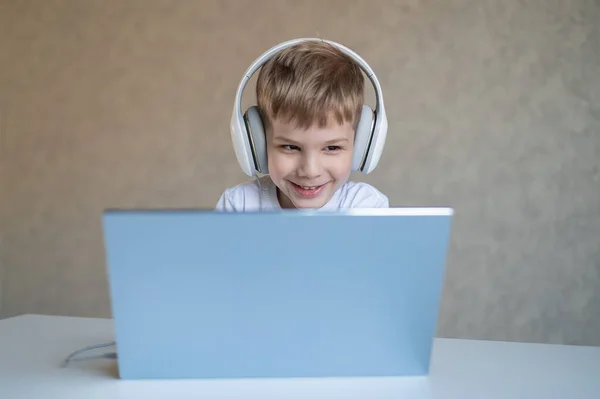 Lindo niño está viendo dibujos animados o una película en un ordenador portátil. Niño caucásico se sienta en una mesa en casa y escucha música en los auriculares. — Foto de Stock