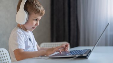 Sevimli çalışkan çocuk klavyede yazı yazıyor ve dizüstü bilgisayara dikkatlice bakıyor. Beyaz bir öğrenci bir masaya oturur, kulaklıkla müzik dinler ve ödev yapar. Uzaklık eğitimi.