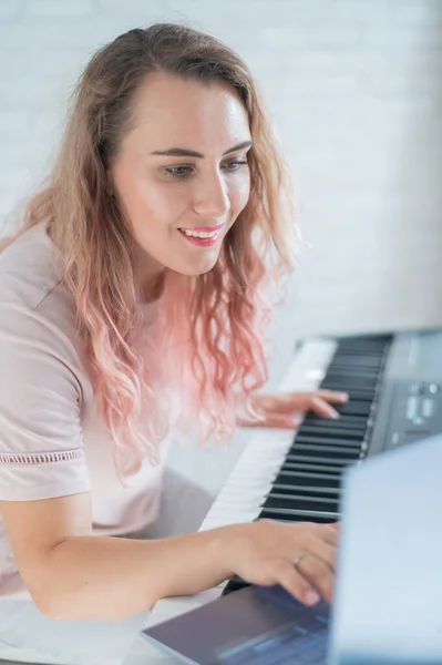 Egy barátságos nő játszik az elektronikus zongorán, és videóblogot vezet a laptopján. Maradj otthon. Hangszertanár. Távolságtartó zene karantén alá helyezve. — Stock Fotó