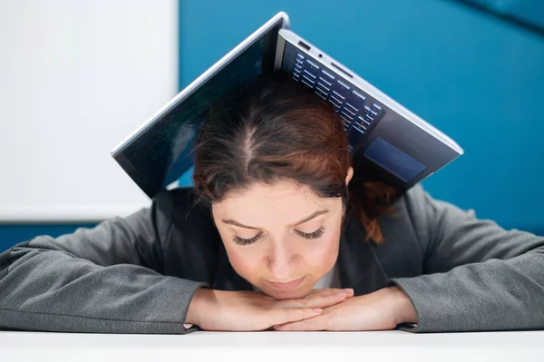 Problemer på jobben. En utslitt kvinne brettet laptopen sin over hodet ved pulten sin. Forretningskvinne i frustrasjon begravd under datamaskinen. Reduksjon og arbeidsledighet. – stockfoto