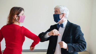 Olgun, gri saçlı bir adam ve kırmızı saçlı, maskeli bir kadın dirseklerini selamlıyor. El sıkışmaya ve sarılmaya bir alternatif. Meslektaşlar aralarındaki mesafeyi koruyorlar. Popüler bir selamlama şekli..