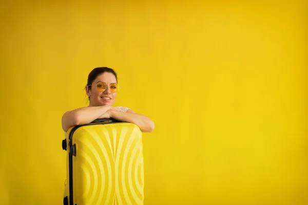 En kvinna med ett perfekt snövitt leende i solglasögon kramar en resväska på en gul bakgrund. Spännande flicka väntar på resa på sommarsemester och snyltar upp till sin väska. — Stockfoto