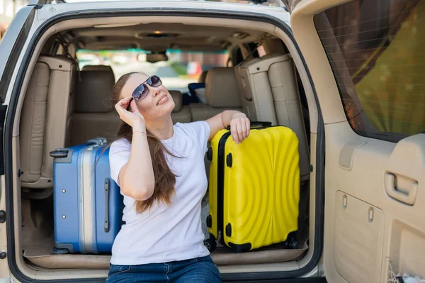 Europäerin mit Sonnenbrille packt ihre Koffer für eine Autofahrt. Schöne glückliche Mädchen sitzt im Kofferraum eines Autos und bereit für die Reise. Gelber Koffer mit Sachen für den Sommerurlaub. — Stockfoto