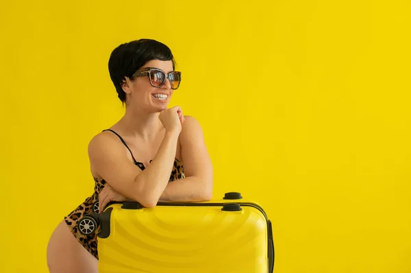 Γυναίκα ποζάρουν σε ένα λεοπαρδαλέ μαγιό και γυαλιά ηλίου με μια βαλίτσα σε κίτρινο φόντο. Μια μελαχρινή με ένα εκθαμβωτικό χαμόγελο σε ένα μονοκίνι πακετάρισε τις αποσκευές της και είναι έτοιμη για διακοπές στην παραλία. — Φωτογραφία Αρχείου