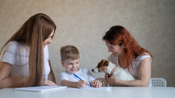 Happy family stays at home. Two women help the boy do school homework. Lesbian couple sitting at the table with their son and a cheerful puppy. Same-sex marriage with a child. Distance learning.