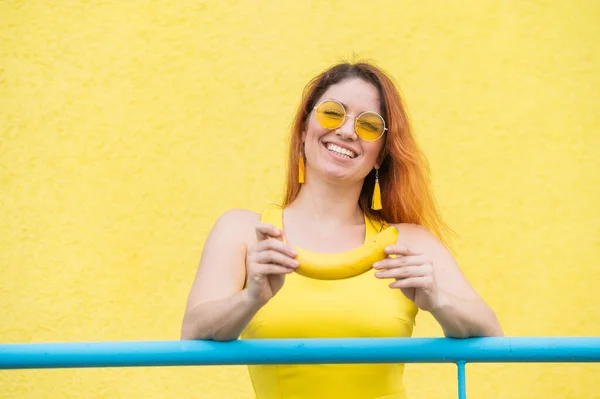 Krásná zrzavá žena ve slunečních brýlích drží banán a blázni kolem. Atraktivní evropská dívka v šatech pózuje na žlutém pozadí půvabně se usmívá. — Stock fotografie
