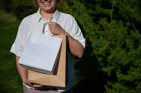 Mulher idosa feliz em óculos detém um pacote ecológico enquanto caminha no parque. Uma senhora idosa encantada fica com compras em sacos de papel ao ar livre . — Fotografia de Stock