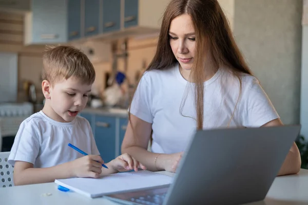 Caucasian woman helps boy learn lessons. The tutor checks the childs homework. Individual home-based quarantine. A diligent boy writes a dictation under the supervision of a teacher.