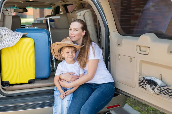 Europäerin sitzt mit ihrem Sohn im Kofferraum eines Geländewagens. Schöne glückliche Mutter liebevoll umarmt Sohn. Gelbes Gepäck für den Sommerurlaub mit dem Auto. Urlaub mit der ganzen Familie. — Stockfoto