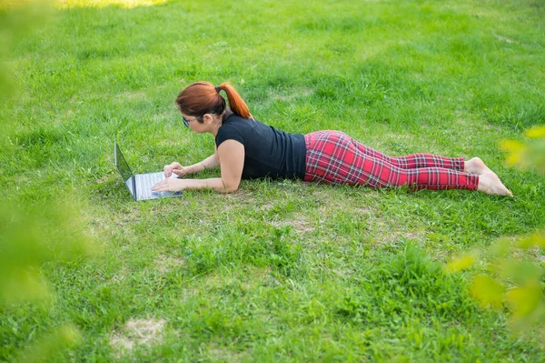 Mulher está comprando em um laptop enquanto deitado em seu estômago na grama verde em um parque. Um estudante com dois copos está estudando em computadores sem fio pessoais enquanto está deitado no gramado. Compras online . — Fotografia de Stock