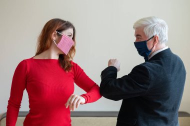 Takım elbiseli olgun, gri saçlı bir adam ve kırmızı elbiseli, tıbbi maske takan kırmızı saçlı bir kadın sosyal mesafeyi koruyup dirseklerini selamlıyorlar. El sıkışmaya ve sarılmaya bir alternatif..
