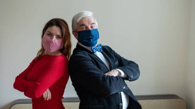 Takım elbiseli olgun gri saçlı bir adam ve kırmızı elbiseli orta yaşlı bir kadın sırt sırta verip tıbbi maskeler takıyorlar. Sosyal mesafeyi koruyan meslektaşların portresi.