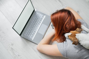 Tecritteki kızıl saçlı kadın yerde bir köpekle yatıyor. Kız sosyal mesafeyi koruyor ve bilgisayarla evden çalışıyor. Bayan öğrenci ev ödevi yapıyor. Koronavirüsün yayılması.