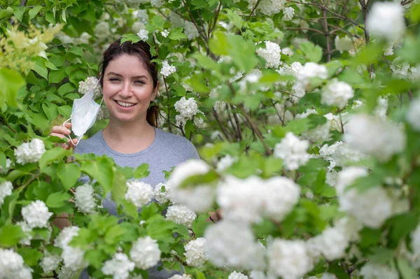 Eine fröhliche rothaarige Frau zieht eine medizinische Maske ab, während sie neben einem blühenden Apfelbaum in einem Park steht. Beendigung der saisonalen allergischen Rhinitis. Das Mädchen wurde von einer Pollenallergie geheilt. — Stockfoto