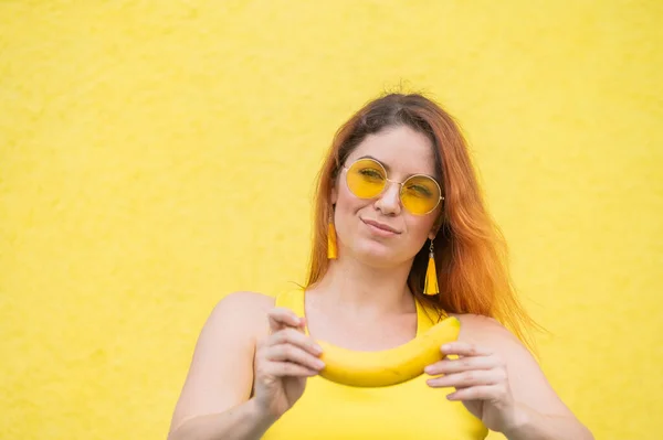 Красивая рыжая женщина в солнечных очках держит в руках банан и дураков. Привлекательная европейская девушка в платье позирует на желтом фоне очаровательно улыбается. — стоковое фото