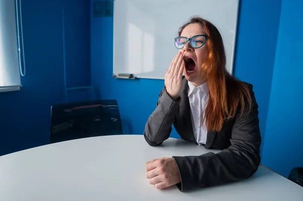 En kvinna i kostym och glasögon gör gäspningar i konferensrummet. En kvinnlig kontorschef med kronisk trötthet sitter vid ett bord i en tråkig verkstad. Partners är sena till ett affärsmöte. — Stockfoto