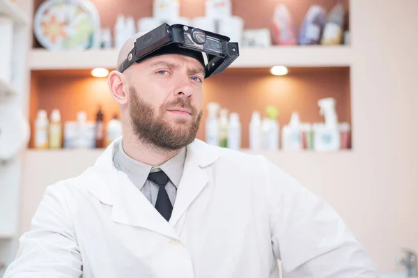 Maschio medico barbuto in un cappotto medico e con una cinghia di ingrandimento w luci seduto sulla scrivania. Attrezzature optometriche. Ufficio medico. — Foto Stock